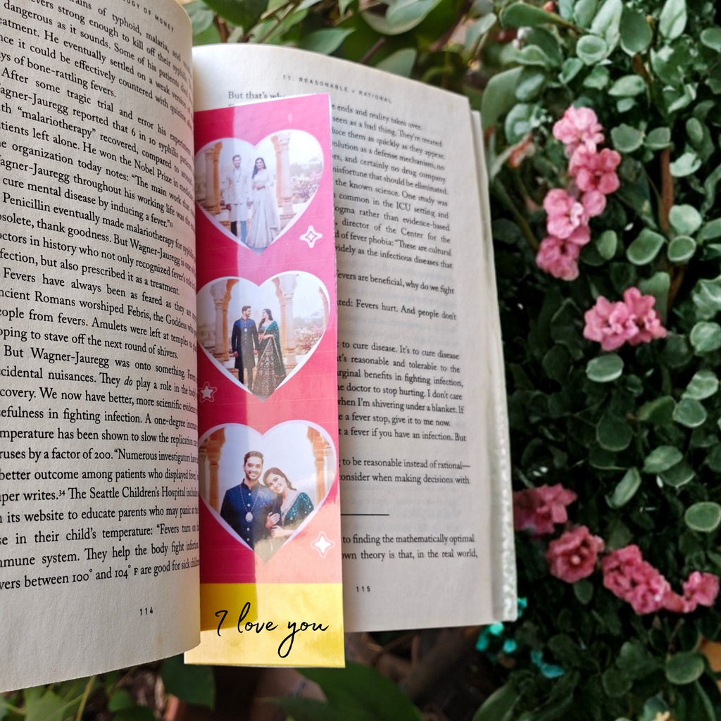 Book with a colorful bookmark featuring heart designs and 'I love you' text, surrounded by greenery and pink flowers.