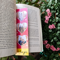 Book with a colorful bookmark featuring heart designs and 'I love you' text, surrounded by greenery and pink flowers.