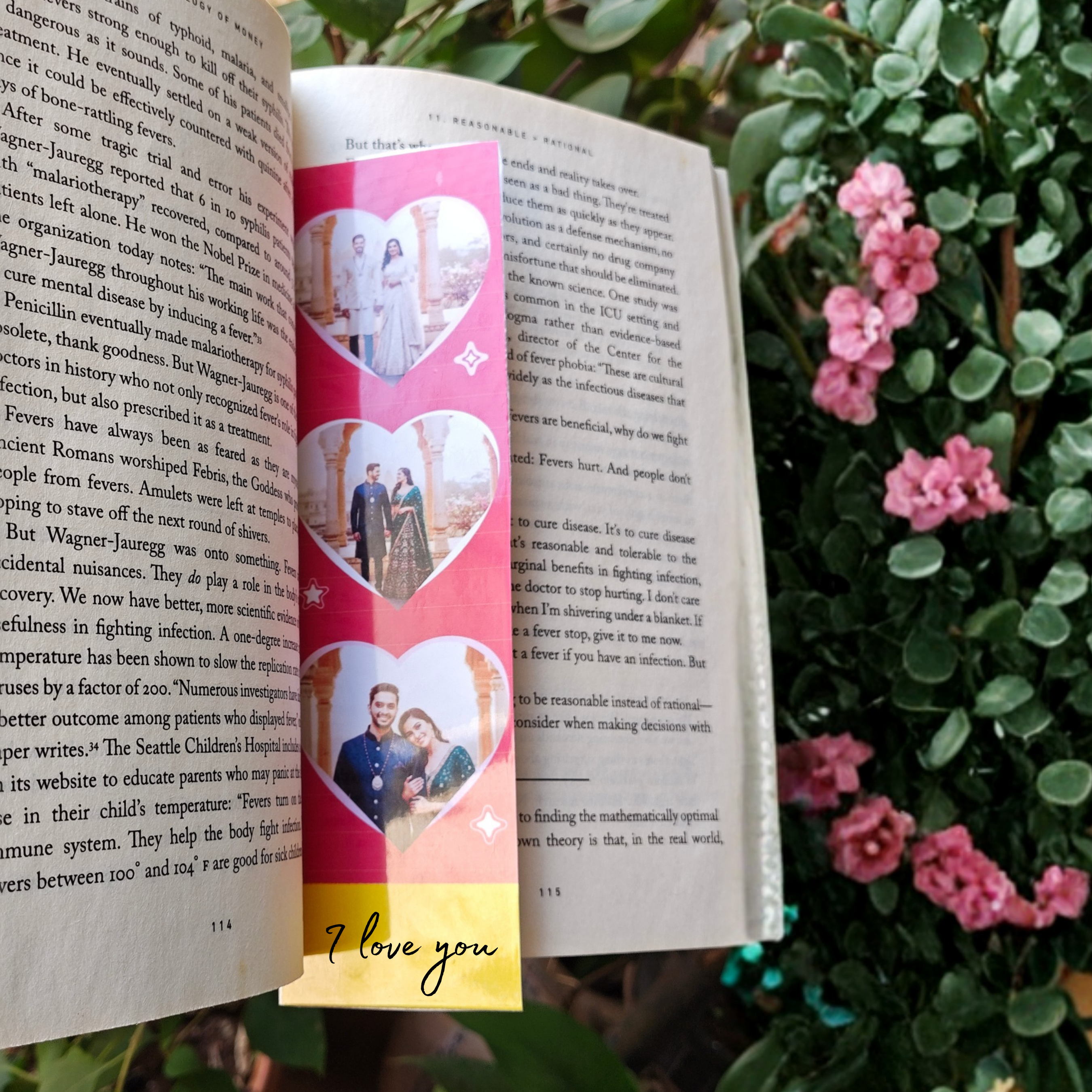 Book with a colorful bookmark featuring heart designs and 'I love you' text, surrounded by greenery and pink flowers.