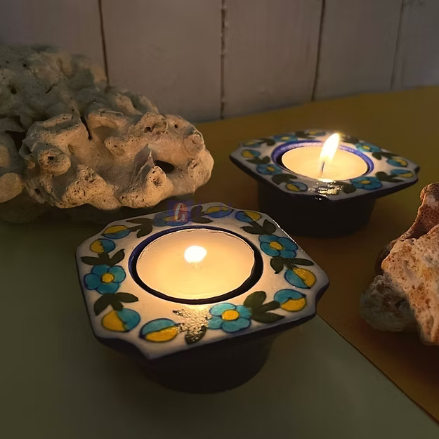 A glowing candle inside a dark blue handcrafted ceramic floral mini tea light holder creating ambient lighting on a tabletop.