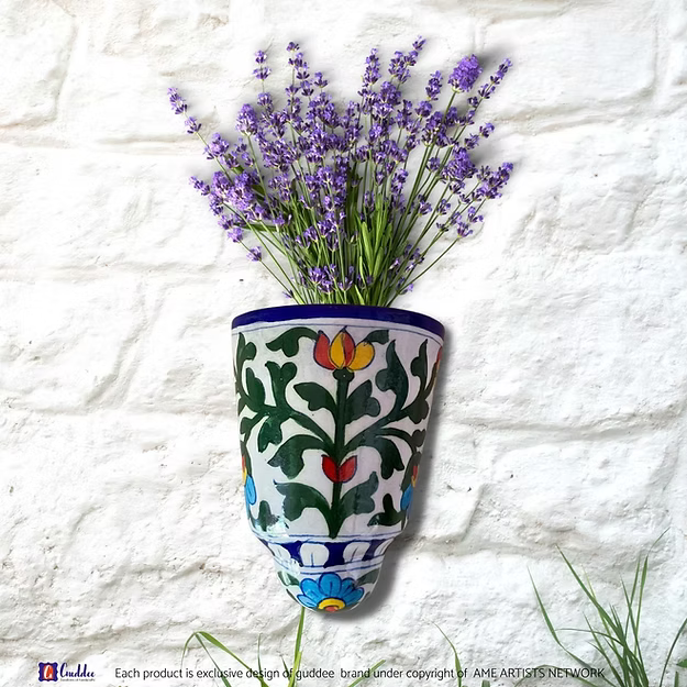 A handcrafted Rangeen Deewana blue pottery wall pocket pot holding fresh purple flower on a modern apartment balcony wall.