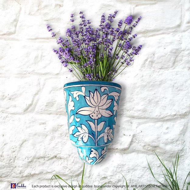 A handcrafted Aasmani blue pottery wall pocket pot holding fresh trailing green vines on a modern apartment balcony wall.