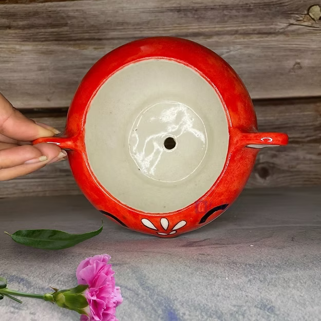 Red ceramic pot with white interior, held by a hand on a wooden surface with a pink flower and green leaf.