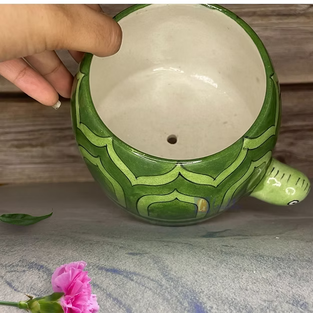 Green ceramic turtle-shaped planter with a hand holding it, on a textured surface with a pink flower.