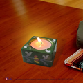 A lit ceramic dark green square candle holder casting a warm ambient glow as a decorative coffee table centerpiece accent.